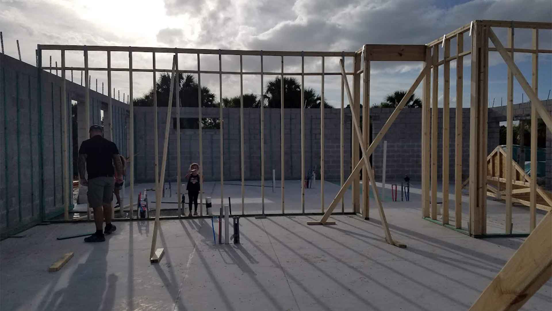 Construction site with incomplete wooden frame and two workers. Partly cloudy sky and concrete ground with scattered materials.